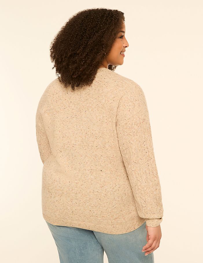 A woman in a textured beige sweater and light blue jeans stands against a plain, light-colored background.