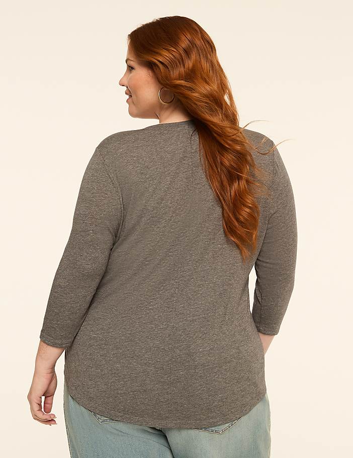 A woman with red hair, wearing a gray shirt and blue jeans, stands against a plain, light-colored background.