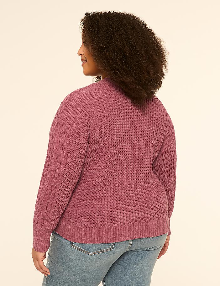 A woman in a textured pink sweater and blue jeans stands against a plain, light-colored background.