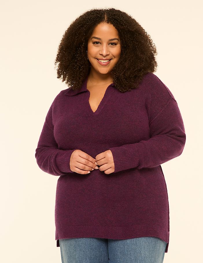 A smiling woman with curly hair wears a purple sweater and jeans against a plain, light background.