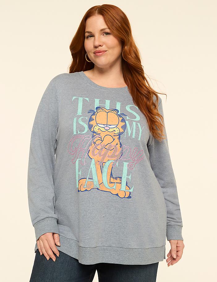 A woman with red hair stands in front of a plain, light-colored background, wearing a blue sweatshirt with a Garfield graphic.