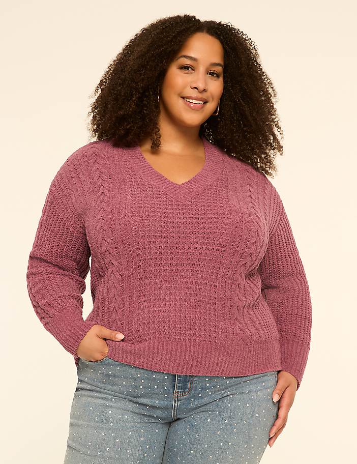 A smiling woman with curly hair in a mauve cable-knit sweater and polka-dot jeans stands against a plain cream background.