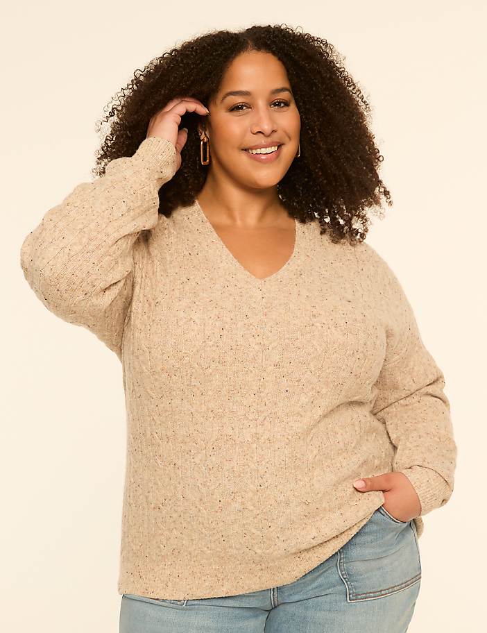 A smiling woman with curly hair in a beige sweater and jeans stands against a plain, light-colored background.