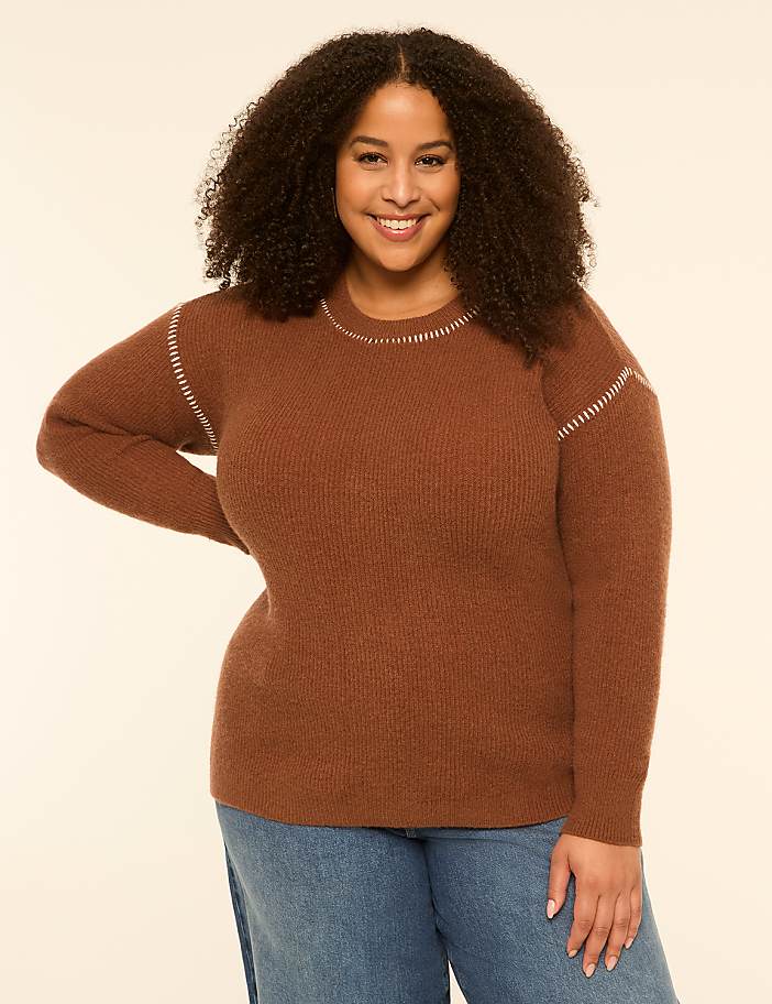 A smiling woman with curly hair wears a brown sweater and blue jeans against a plain, light-colored background.
