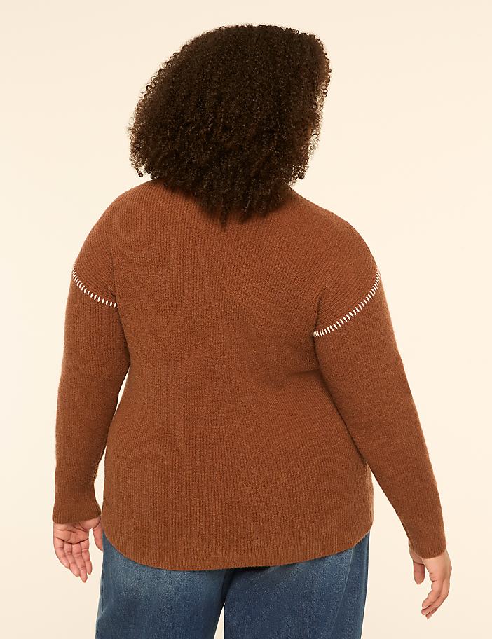 A person wearing a brown sweater and blue jeans stands against a plain, light-colored background.
