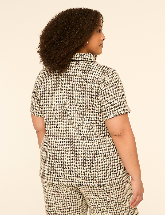 A woman with curly hair, seen from the back, wears a houndstooth patterned shirt and pants against a plain, light-colored background.