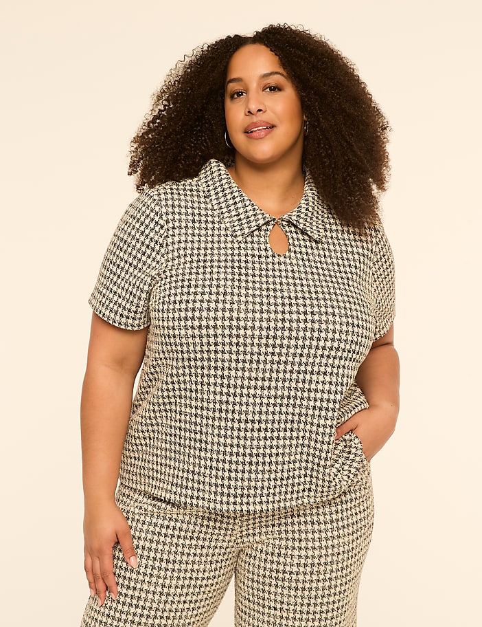 A woman with curly hair stands in the foreground wearing a houndstooth top and pants against a plain, light-colored background.