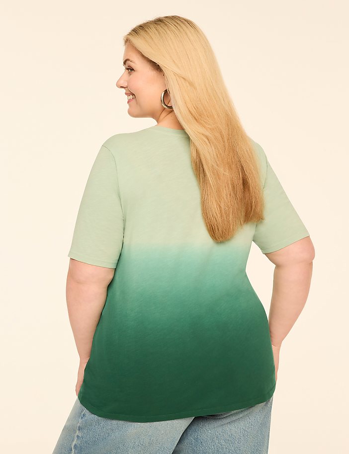 A woman with blonde hair, wearing a green ombre t-shirt and blue jeans, smiles as she stands against a plain, light-colored background.