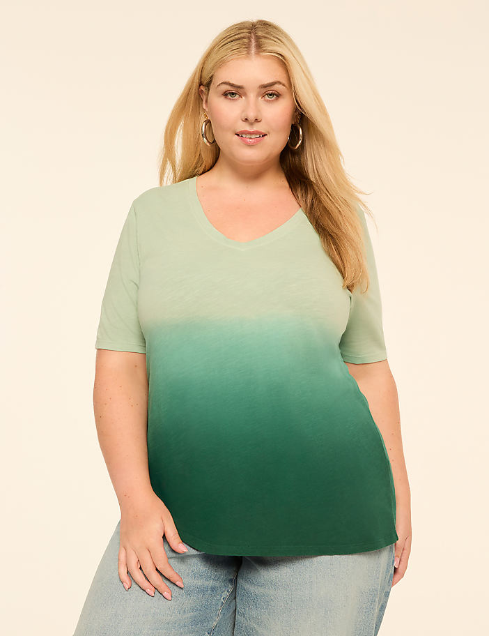 A woman in a light green to dark green ombre t-shirt and jeans stands against a plain, light-colored background.