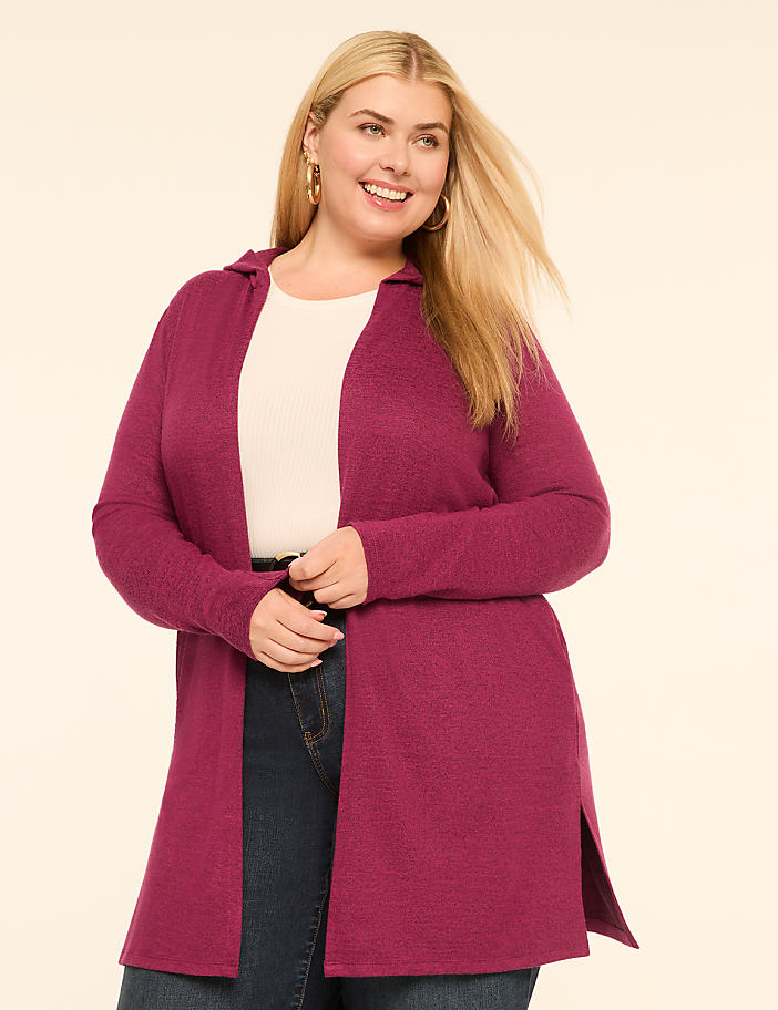 A smiling woman in a magenta cardigan and jeans stands against a plain, light-colored background.