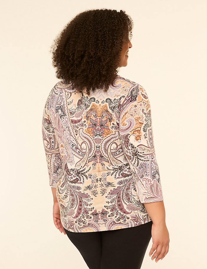 A woman with curly brown hair, wearing a paisley-patterned shirt and black pants, stands against a plain, light-colored background.