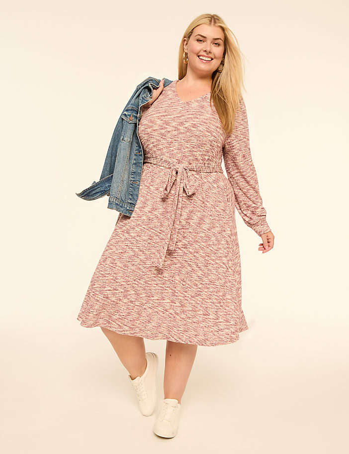 A smiling woman in a pink patterned dress and white sneakers, with a denim jacket slung over her shoulder, poses against a plain cream background.