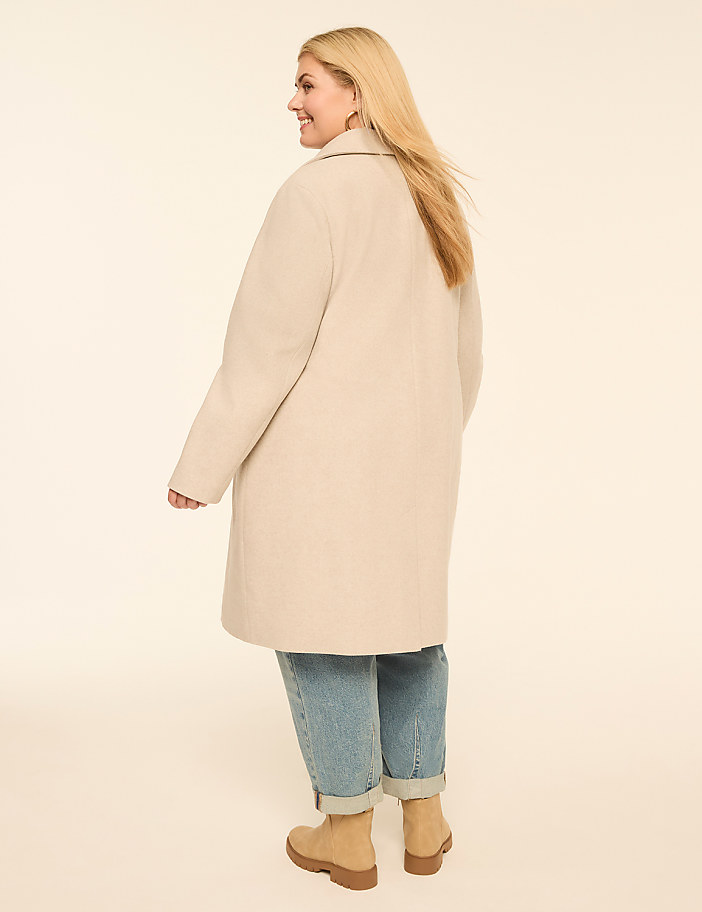 A woman in a cream coat and cuffed jeans stands against a plain, light background.
