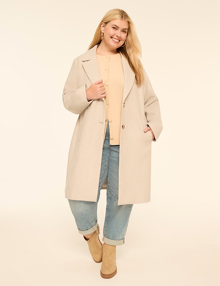 A smiling woman in a beige coat and light blue jeans stands against a plain cream background.