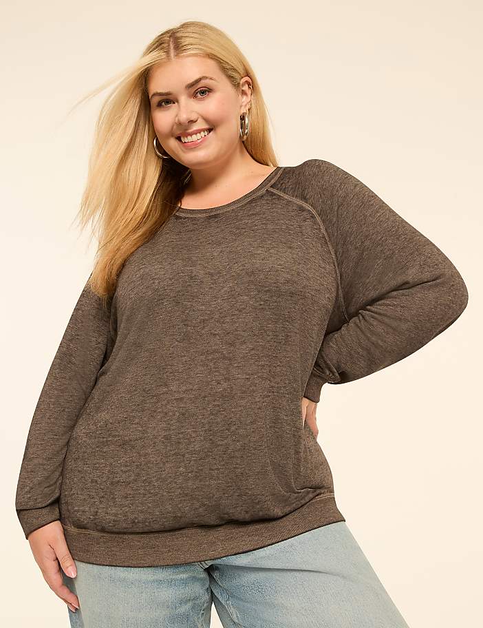 A smiling woman with blonde hair and hoop earrings poses in a brown long-sleeved shirt and light blue jeans against a plain, light-colored background.