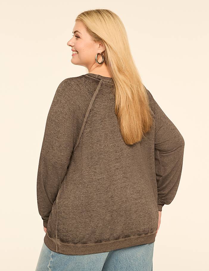 A woman with blonde hair wearing a brown sweater and blue jeans stands against a plain, light-colored background.