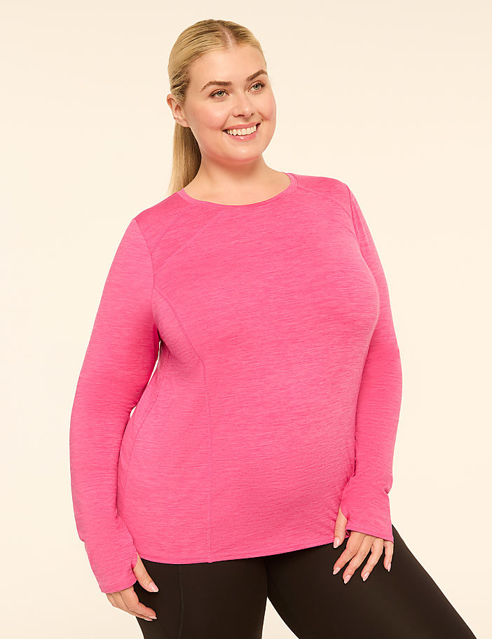 A smiling woman in a pink long-sleeved shirt and black leggings stands against a plain, light-colored background.