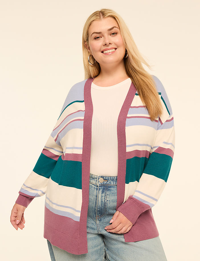 A smiling woman with blonde hair wears a striped cardigan over a white top and jeans, with a plain, light-colored background.