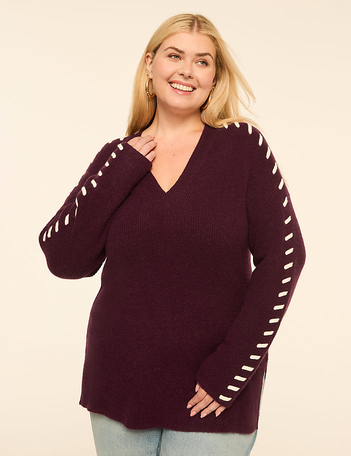 A smiling woman in a plum v-neck sweater with white stitching on the sleeves stands against a plain cream background.