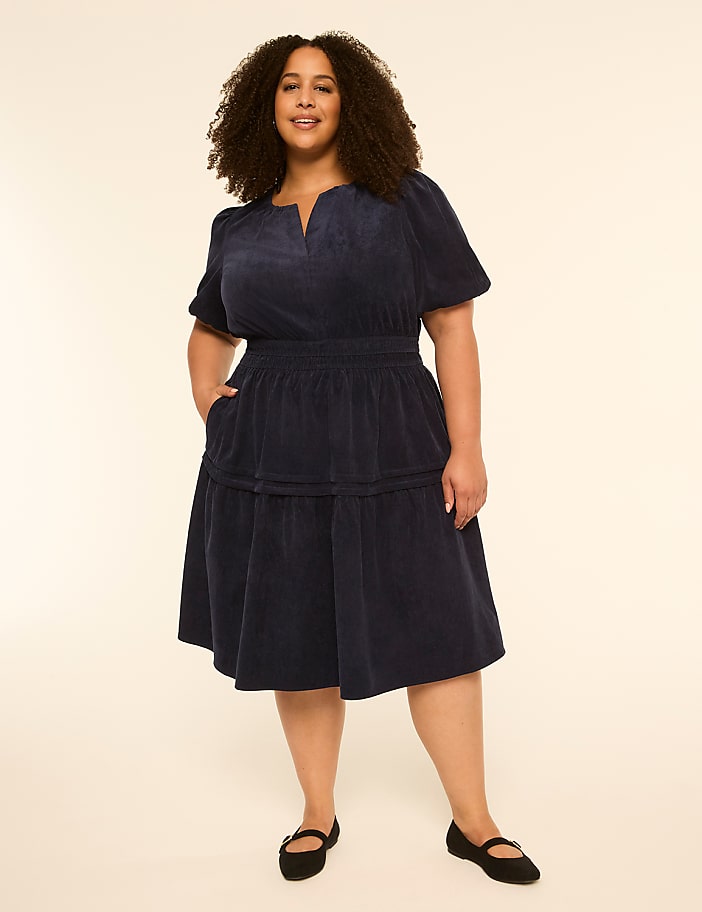 A woman in a navy corduroy dress stands against a plain, light-colored background.