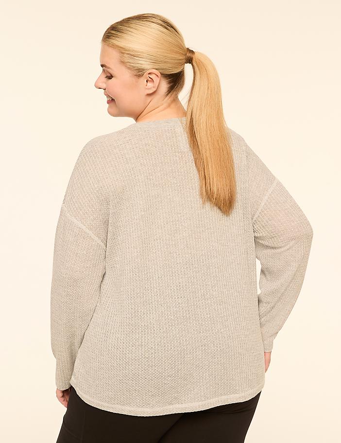 A woman with blonde hair in a ponytail, wearing a light gray sweater, stands against a plain, light-colored background.