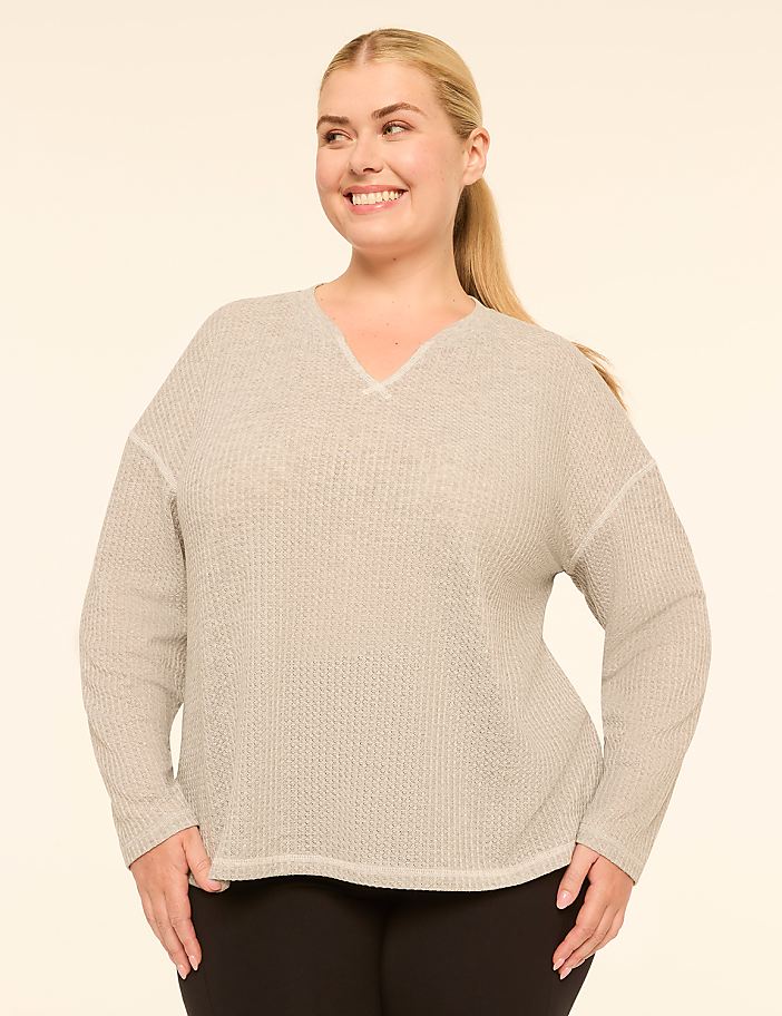 A smiling woman with blonde hair pulled back in a ponytail poses in a light gray, textured v-neck sweater and black pants against a plain, light beige background.