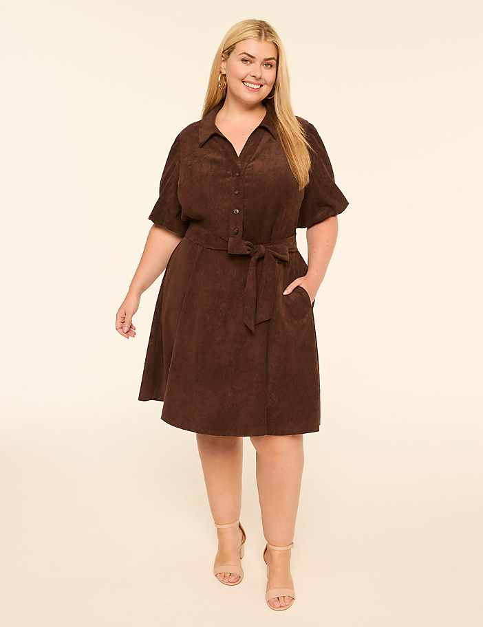 A smiling woman in a brown corduroy dress stands against a plain, light-colored background.