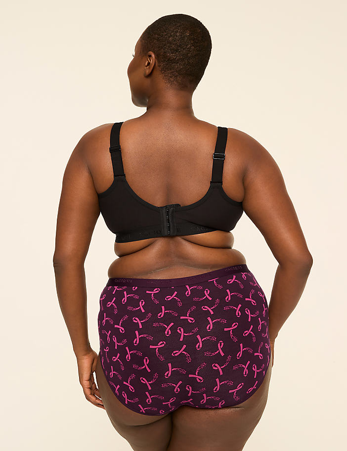 A woman from behind, wearing a black bra and purple patterned underwear, stands against a plain, light-colored background.