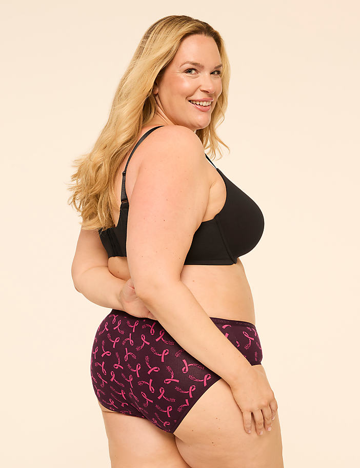 A woman in a black bra and patterned underwear smiles over her shoulder against a plain background.