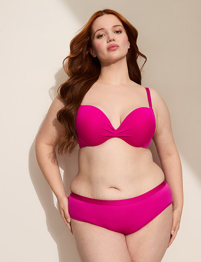 A woman with long, wavy red hair poses in a bright pink strapless bra and matching bottoms against a plain, light-colored wall with a shadow cast from the left.