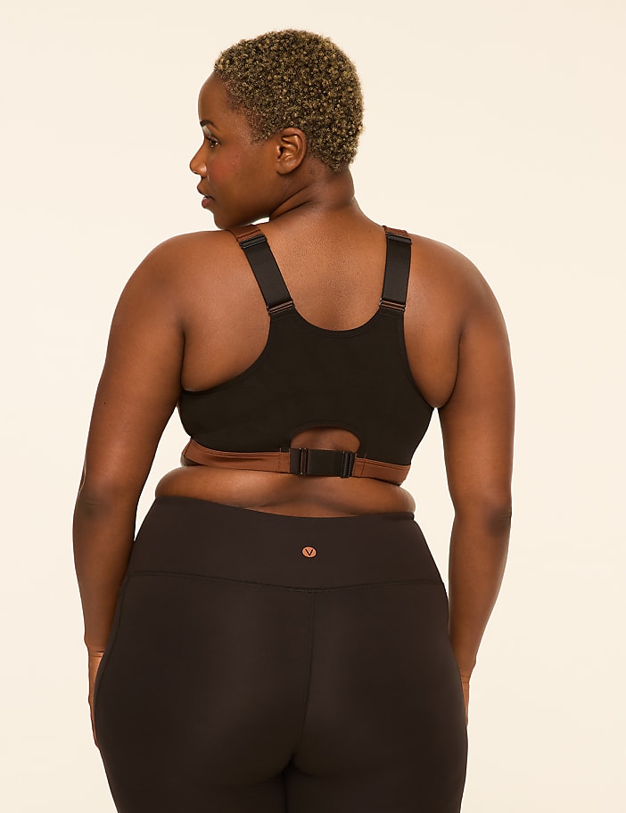 A woman with short, curly hair faces away from the camera, wearing a black sports bra and black leggings against a plain, light-colored background.