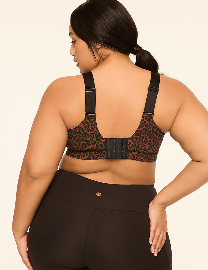 A woman with dark hair pulled back in a ponytail, wearing a leopard-print sports bra and black leggings, stands against a plain, light-colored background.