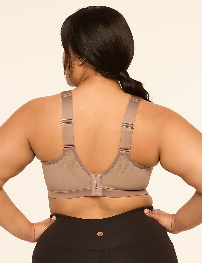 A woman with dark hair and a tan sports bra stands with her hands on her hips against a plain beige background.