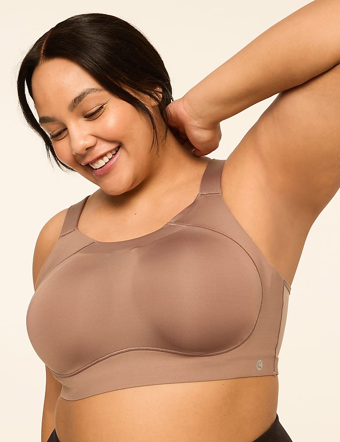 A smiling woman in a taupe sports bra with her arm raised against a plain, light background.