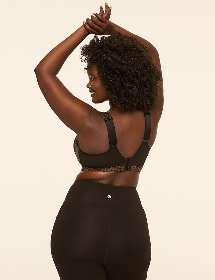 A woman with dark curly hair and dark skin, wearing a leopard print sports bra and black leggings, poses with her arms raised against a plain, light-colored background.