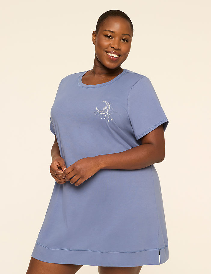 A smiling woman in a blue nightgown with a celestial design stands against a plain, light-colored background.