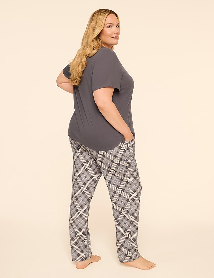 A woman in a grey t-shirt and plaid pajama pants stands with her hands in her pockets against a plain, light-colored background.