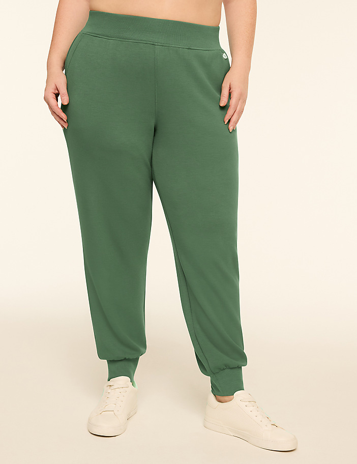 A person wearing green sweatpants and white sneakers stands against a plain, light-colored background.