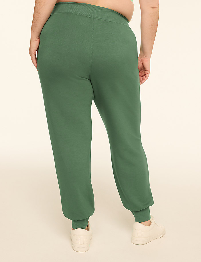 A person wearing green sweatpants and white sneakers stands against a plain, light-colored background.