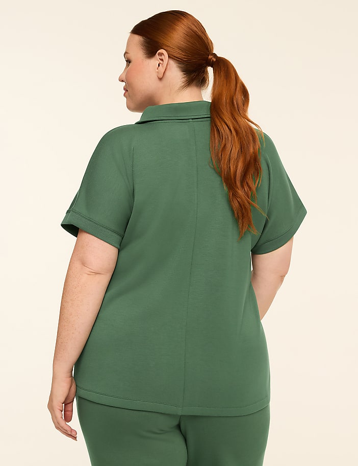 A woman with auburn hair pulled into a ponytail stands facing away from the camera, wearing a green polo shirt and pants against a plain, light-colored background.