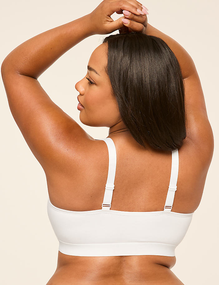 A woman with dark hair and brown skin, wearing a white bralette, poses with her arms raised behind her head against a plain, light-colored background.