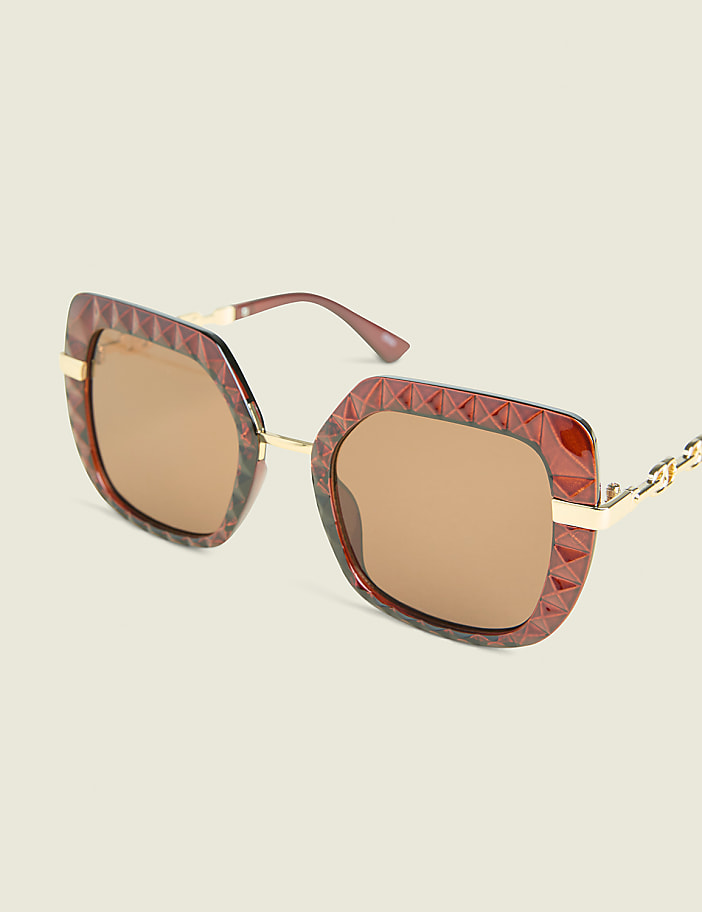 A pair of oversized, square, brown-patterned sunglasses with gold accents rests on a plain, light-colored background.