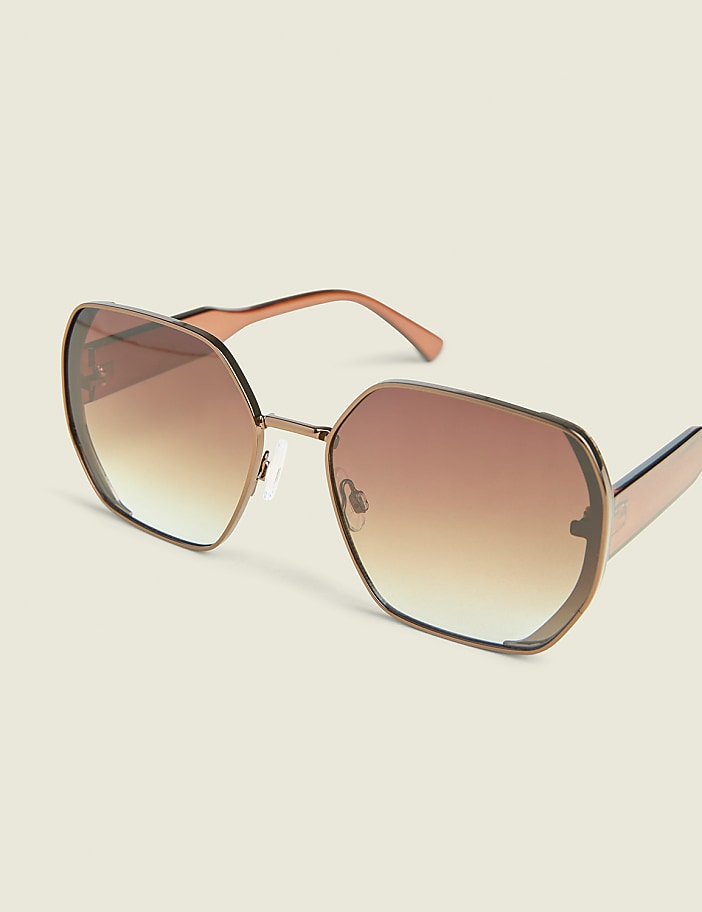 A pair of hexagonal, rose gold sunglasses with gradient brown lenses rests on a plain, light beige background.