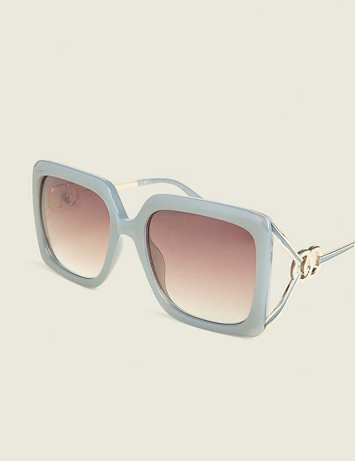A pair of light blue, oversized square sunglasses with gold accents rests on a pale yellow background.