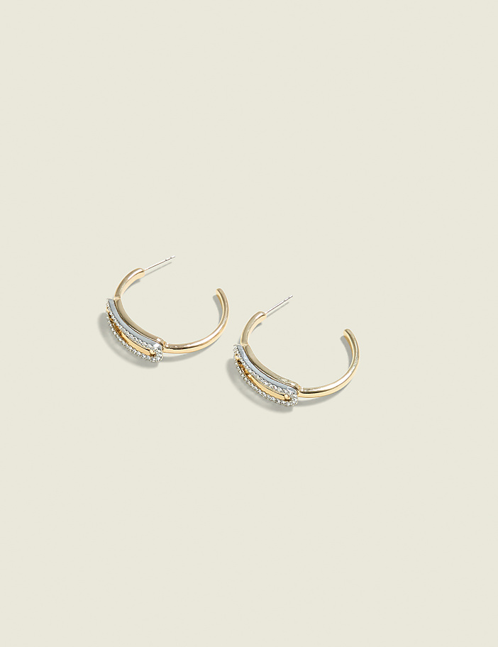A pair of gold hoop earrings with a textured band of small stones rests on a pale, textured background.