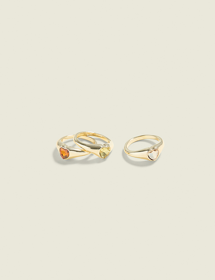Three gold rings with heart-shaped gemstones are arranged on a plain, light-colored background.