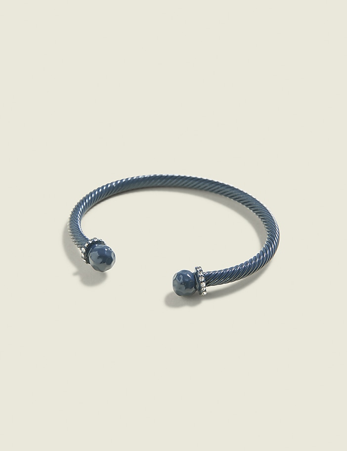 A dark blue, twisted metal cuff bracelet with blue stone ends and small diamonds rests on a pale, textured background.