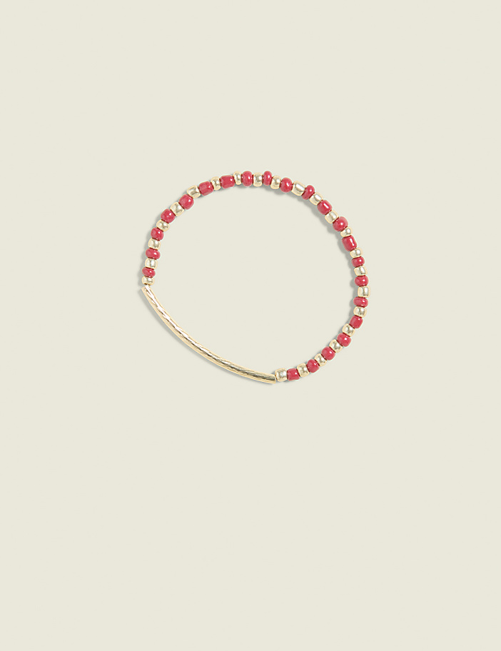 A delicate gold bar bracelet with red beads is centered against a soft, pale yellow background.