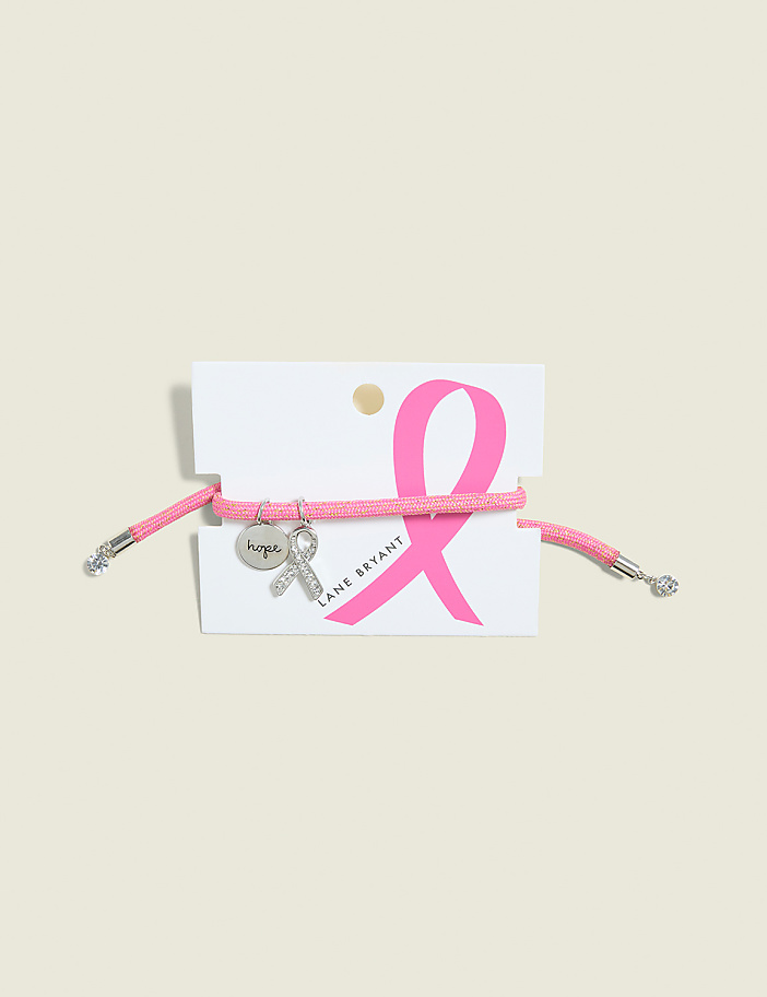 A pink cord bracelet with a silver charm hangs on a white card featuring a pink breast cancer ribbon and the Lane Bryant logo, set against a pale, textured background.