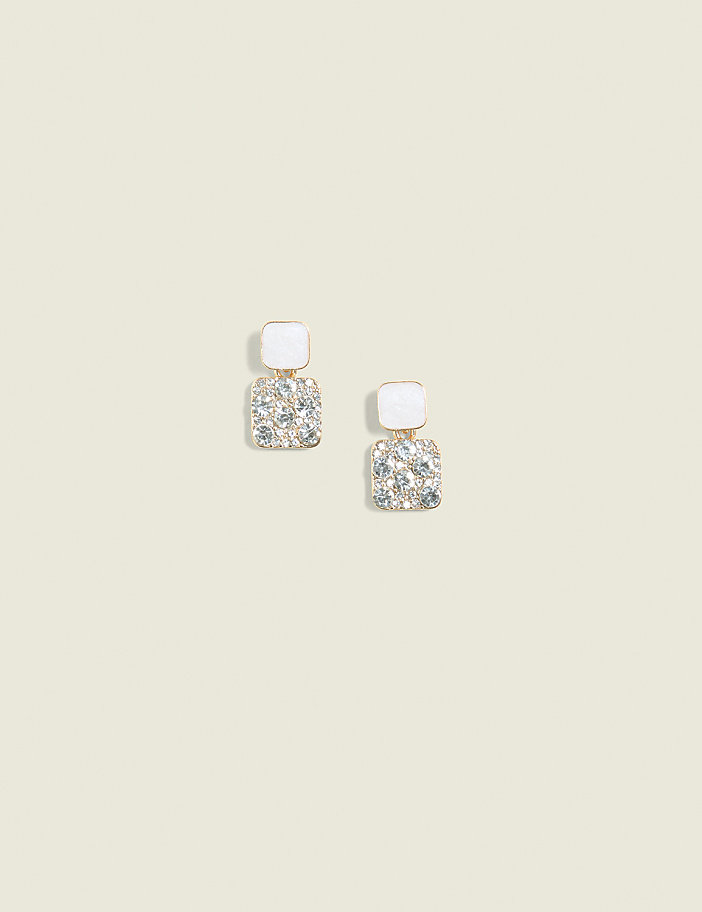A pair of square, white-topped earrings with sparkling rhinestones dangle against a plain, light beige background.